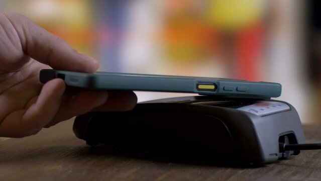 Male's hand puts phone on payment terminal to transfer money for purchase. Convenient shopping. Blurred background. Time efficiency concept