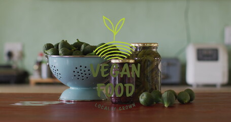 A bowl and jar labeled VEGAN FOOD sit on table surrounded by green fruits