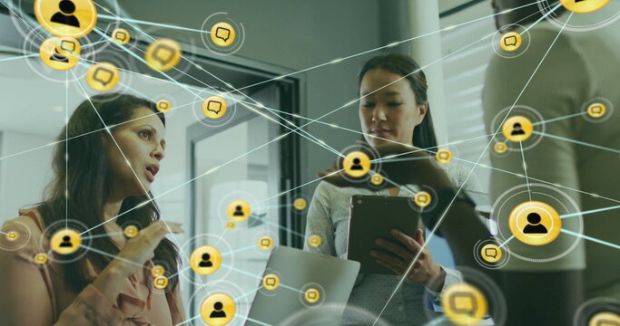 Image of connected icons over diverse female coworkers discussing reports in office