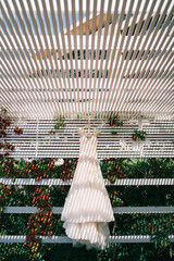 Wedding dress hangs on a hanger in the striped shade of a wooden canopy in the garden