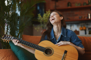 Happy woman playing acoustic guitar and laughing while sitting on a couch in cozy living room