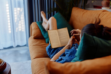 Woman relaxing on a couch by the window, engrossed in a book in her cozy living room at home