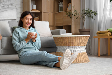 Beautiful young Asian woman in stylish hoodie with cup of tea sitting on floor at home