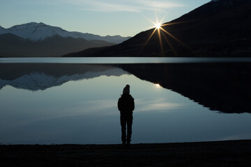 Silhouette woman standing alone looking at a reflection of mountains at the shore of a lagoon,...