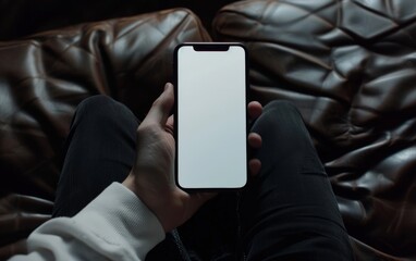 Mockup of a blank iPhone screen sitting on the lap, top view of a hand holding a phone with a white screen, wearing black pants and shirt with a dark background.