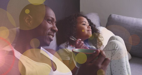 Image of dots moving over happy african american father and daughter eating and watching tv