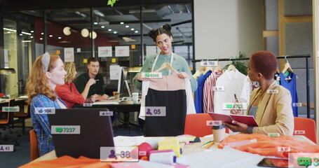 Anmation of social media icons over diverse team of fashion designers discussing together at office