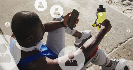 Image of profile icons over tired african american athlete sitting and looking at smartphone