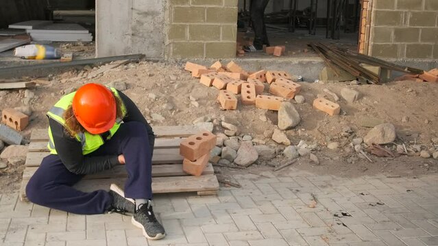 Unfortunate worker in an orange helmet broke his leg when falling from the scaffolding of an unfinished building needs the help of employees and call an ambulance.