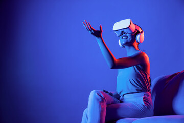 Smart female sitting on sofa surrounded by neon light wear VR headset connect metaverse, futuristic...