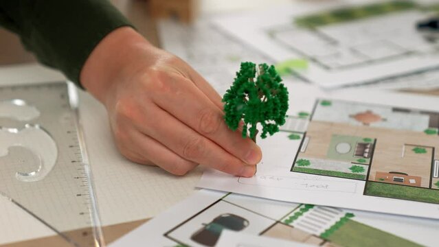 A video of architect using tree and home model to measure blueprint. Architect designing house construction on a table at studio, with architectural plan scattered around. Focus on hand. Delineation.