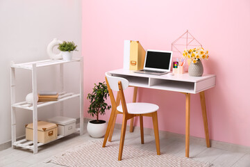 Shelving unit and workplace with bouquet of narcissus flowers, stationery and laptop near pink wall...