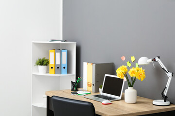Shelving unit and workplace with bouquet of narcissus flowers, lamp and laptop near black wall in stylish office