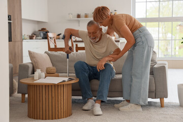 Mature woman helping her husband with crutch to stand up from sofa at home