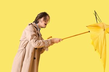 Young woman with broken umbrella and strong wind on yellow background