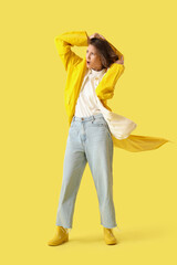 Young woman in raincoat with strong wind on yellow background