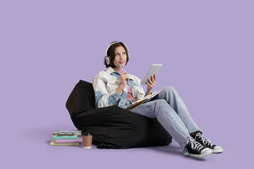 Female student in headphones with tablet computer and copybooks studying on beanbag against lilac...