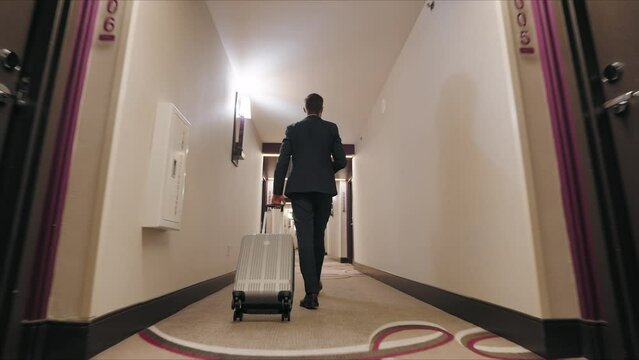 Businessman in a sleek, dark suit walks down a hotel corridor, coffee in one hand and a suitcase in another. His brisk, determined steps showcase his mastery of balancing travel and tasks. 