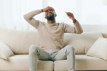 A tired African American man sitting on a couch at home, feeling stressed and frustrated due to a...