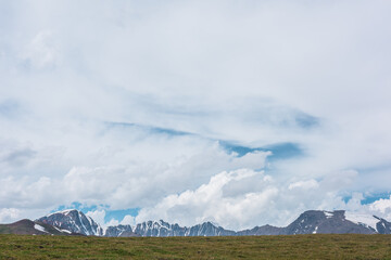 Scenic minimalist alpine view from green meadow to big snowy rocky ridge under clouds in blue sky....