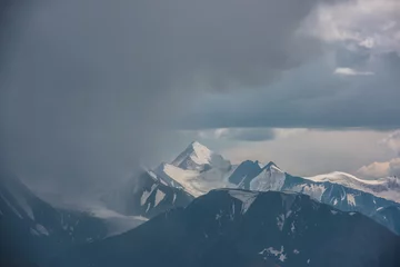 Fototapeten Dark atmospheric landscape with mountain silhouettes and large snow-capped peaked top in rainy low clouds. Dramatic alpine view to high mountains in gray cloudy sky. Big snowy mountain range in rain. © Daniil