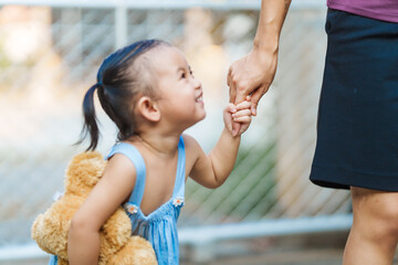 young mom shares a tender moment with her playful, happy Asian two-year-old female toddler, as they explore outdoors, hand in hand, accompanied by her cherished brown teddy bear.