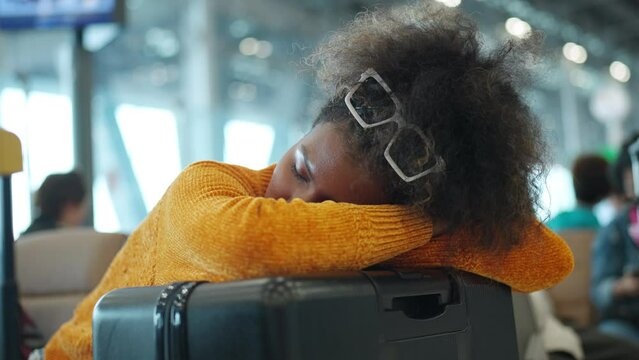 African woman passenger with luggage napping on passenger seat during waiting flight boarding airport terminal departure hall. Attractive girl enjoy travel on holiday vacation with air transportation.