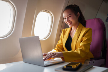 Young Asian business woman in yellow suit uses laptop sitting near window on airplane to do online...
