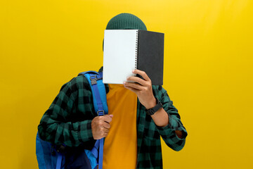 A young Asian man, dressed in a beanie hat and casual shirt and carrying a backpack, is covering...