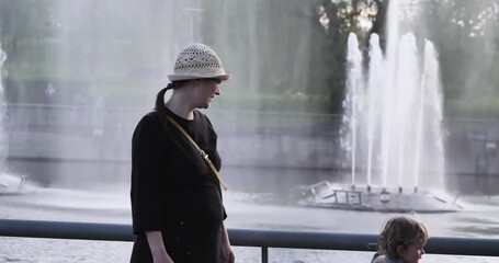 Pretty girl in dress spreads her arms from seeing. Woman in hat and black dress is waving her hands. Surprised by beauty of fountain under rays of setting sun. Concept: Woman is surprised by fountain