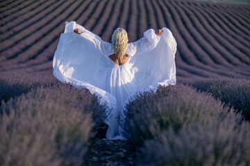 Blonde woman poses in lavender field at sunset. Happy woman in white dress holds lavender bouquet....