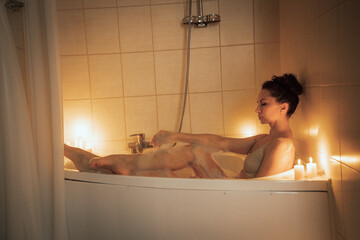 A woman is sitting in a bathtub with candles lit around her. Scene is relaxing and calming.
