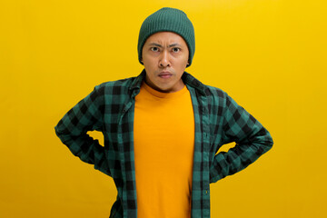 Angry young Asian man, wearing a beanie hat and casual outfit, stands with arms akimbo, looking furious at the camera. Isolated on a yellow background