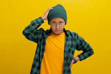 Anxious young Asian man, dressed in a beanie hat and casual shirt, panics and grabs his head, visibly worried and frustrated. He looks at the camera with a realization of a significant failure.