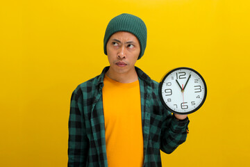An uncertain Asian man, dressed in a beanie hat and casual shirt, holds a clock, appearing confused...
