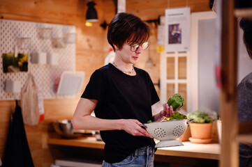 Fototapeta premium This image portrays a young adult focused on preparing fresh vegetables in a rustic kitchen. The scene is warmly lit