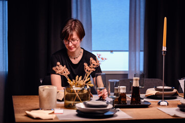 Fototapeta na wymiar Young adult carefully arranging a floral centerpiece at a sophisticated dining table. The scene is set with stylish tableware and mood lighting, capturing a moment of preparation for a dinner party. 