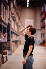 Captured in a spacious warehouse, young woman in casual attire, reaching for an item on a high...