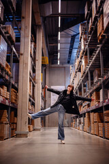 Captured in a dynamic pose, a young woman playfully dancing in the aisle of a warehouse. Her spirited movement, highlighted by a stylish black puffer jacket and jeans, 