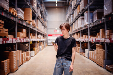 Young woman engaging with her smartphone while standing in a warehouse aisle. Her focused expression and casual style, complete with glasses and modern attire