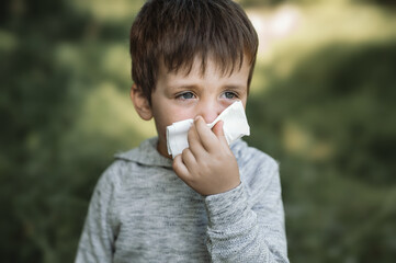 A little boy 4 years old blows his nose into a paper napkin against a background of greenery. Spring exacerbation of allergy to poplar fluff. Allergies in children hay fever