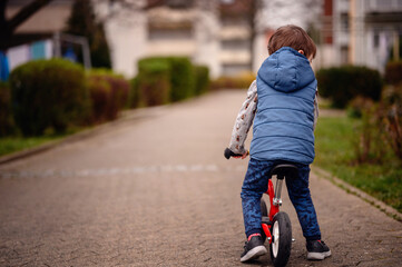 Captured from behind, a young boy pushes his red scooter along a paved path in a quiet suburban area. He is dressed warmly in a blue vest and patterned long-sleeve shirt, 