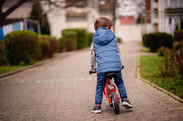 Captured from behind, a young boy pushes his red scooter along a paved path in a quiet suburban...