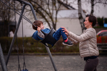 Obraz premium A heartwarming moment of a mother pushing her young son on a swing at a local park. The boy, laughing with glee, reaches out his feet to meet his mother's hands in a playful game. 