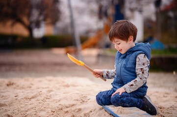 A young boy sits in a sandbox, deeply engaged in play. He holds a sandbox toy, lost in his creative...