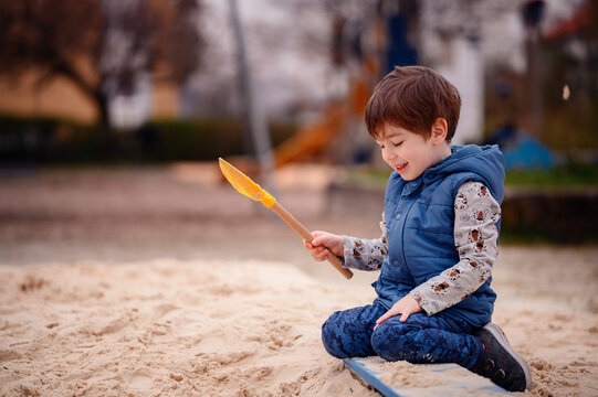A young boy sits in a sandbox, deeply engaged in play. He holds a sandbox toy, lost in his creative world. His focused expression and the soft backdrop of the playground emphasize a peaceful