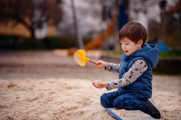 A young boy sits in a sandbox, deeply engaged in play. He holds a sandbox toy, lost in his creative...