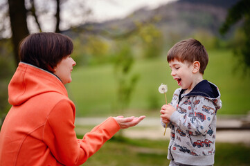 A tender moment in a lush green park, this photo captures a young boy blowing dandelion seeds while his mother holds it gently for him. 