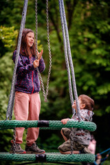 A young girl in a polka-dotted jacket clutches a chain swing, looking upwards with joy, while a...