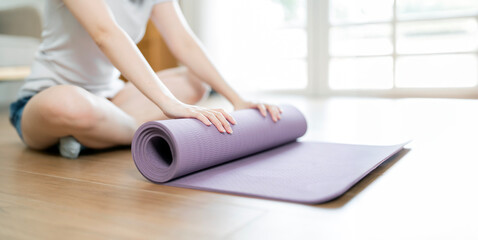 Young yoga Woman rolling her lilac mat after a yoga, sitting on wooden floor near a window,